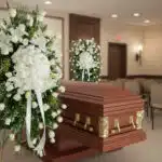 Ataúd de madera pulida con herrajes dorados junto a coronas de flores blancas en una sala de velatorio