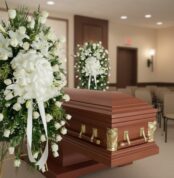 Ataúd de madera pulida con herrajes dorados junto a coronas de flores blancas en una sala de velatorio