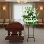 Arreglo floral funerario de lirios blancos y hortensias sobre pedestal negro al lado de un ataúd de madera oscura en una sala de velatorio