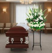 Arreglo floral funerario de lirios blancos y hortensias sobre pedestal negro al lado de un ataúd de madera oscura en una sala de velatorio