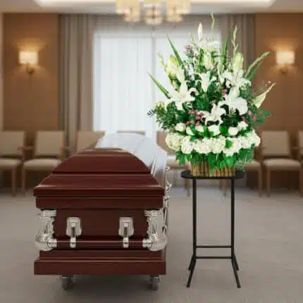Arreglo floral funerario de lirios blancos y hortensias sobre pedestal negro al lado de un ataúd de madera oscura en una sala de velatorio