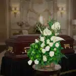 Arreglo de flores blancas con rosas y anturios sobre mesa redonda, colocado frente a un ataúd de madera oscura con herrajes dorados en una sala funeraria elegante