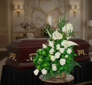 Arreglo de flores blancas con rosas y anturios sobre mesa redonda, colocado frente a un ataúd de madera oscura con herrajes dorados en una sala funeraria elegante