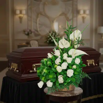 Arreglo de flores blancas con rosas y anturios sobre mesa redonda, colocado frente a un ataúd de madera oscura con herrajes dorados en una sala funeraria elegante