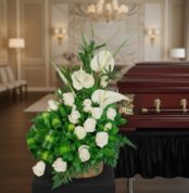 Arreglo de flores blancas con rosas y anturios sobre base de madera al lado de un ataúd de madera barnizada en una elegante sala de velación