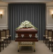 Ataúd de madera barnizada con adornos metálicos plateados y arreglo de flores blancas sobre la tapa, situado en el centro de una sala de velatorio con filas de sillas y cortina gris de fondo