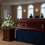 Ataúd de madera barnizada sobre mesa cubierta con tela azul en una sala de velatorio, junto a un pedestal con arreglo de flores blancas y tres personas de pie al fondo