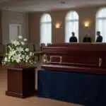 Ataúd de madera barnizada sobre mesa cubierta con tela azul en una sala de velatorio, junto a un pedestal con arreglo de flores blancas y tres personas de pie al fondo