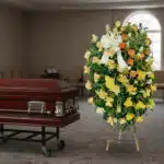 Corona de flores amarillas y naranjas con lazo blanco junto a un ataúd de madera barnizada en una sala funeraria