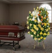 Corona de flores amarillas y naranjas con lazo blanco junto a un ataúd de madera barnizada en una sala funeraria