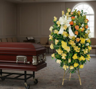 Corona de flores amarillas y naranjas con lazo blanco junto a un ataúd de madera barnizada en una sala funeraria