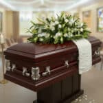Ataúd de madera barnizada color caoba con herrajes plateados y arreglo de flores blancas en la parte superior, situado en una sala velatoria iluminada