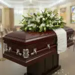 Ataúd de madera barnizada color caoba con herrajes plateados y arreglo de flores blancas en la parte superior, situado en una sala velatoria iluminada