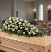 Centro de flores blancas con rosas y follaje verde colocado sobre un ataúd de madera clara en una sala de velatorio