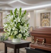 Arreglo de flores blancas con lirios y rosas en maceta blanca sobre mesa, junto a ataúd de madera pulida en una sala velatoria