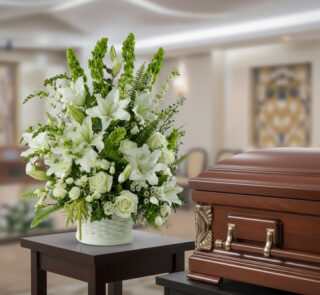 Arreglo de flores blancas con lirios y rosas en maceta blanca sobre mesa, junto a ataúd de madera pulida en una sala velatoria