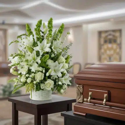 Arreglo de flores blancas con lirios y rosas en maceta blanca sobre mesa, junto a ataúd de madera pulida en una sala velatoria