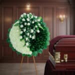 Arreglo floral fúnebre redondo en tonos blanco y verde sobre caballete de madera, colocado junto a un ataúd de madera barnizada en una sala velatoria