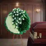 Arreglo floral fúnebre redondo en tonos blanco y verde sobre caballete de madera, colocado junto a un ataúd de madera barnizada en una sala velatoria