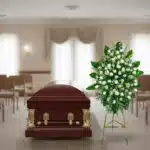 Ataúd de madera barnizada con herrajes dorados junto a un arreglo floral blanco y verde en una sala de velatorio iluminada