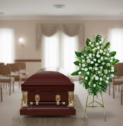 Ataúd de madera barnizada con herrajes dorados junto a un arreglo floral blanco y verde en una sala de velatorio iluminada