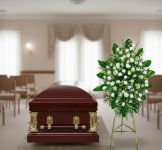 Ataúd de madera barnizada con herrajes dorados junto a un arreglo floral blanco y verde en una sala de velatorio iluminada