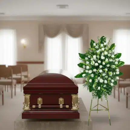 Ataúd de madera barnizada con herrajes dorados junto a un arreglo floral blanco y verde en una sala de velatorio iluminada