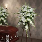 Arreglo floral grande de flores blancas sobre caballete de madera junto a un ataúd de madera barnizada en una sala funeraria