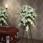 Arreglo floral grande de flores blancas sobre caballete de madera junto a un ataúd de madera barnizada en una sala funeraria
