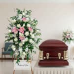 Arreglo floral alto con lirios rosas y flores blancas al lado de un ataúd de madera barnizada en una sala velatoria
