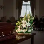 Arreglo de flores blancas y verdes sobre mesa de madera junto a un ataúd marrón en una sala de velatorio con personas sentadas al fondo