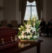 Arreglo de flores blancas y verdes sobre mesa de madera junto a un ataúd marrón en una sala de velatorio con personas sentadas al fondo