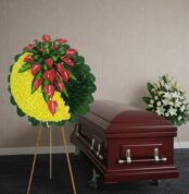 Corona funeraria redonda con flores amarillas y anturios rojos sobre caballete al lado de un ataúd de madera barnizada en una sala velatoria