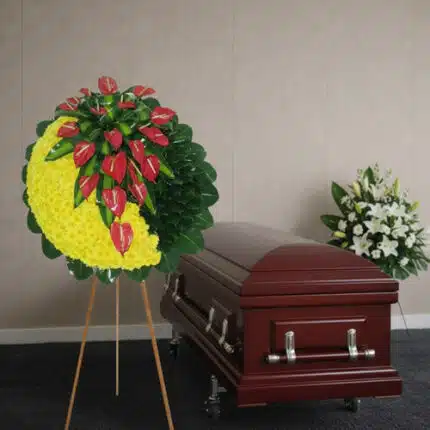 Corona funeraria redonda con flores amarillas y anturios rojos sobre caballete al lado de un ataúd de madera barnizada en una sala velatoria