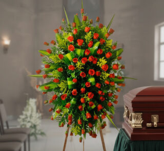 Corona funeraria grande de rosas naranjas y hojas verdes sobre caballete junto a un ataúd de madera en una sala velatoria