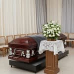 Ataúd de madera barnizada con herrajes plateados en una sala de velatorio, junto a un pedestal con mantel de encaje y un arreglo de flores blancas