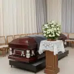 Ataúd de madera barnizada con herrajes plateados en una sala de velatorio, junto a un pedestal con mantel de encaje y un arreglo de flores blancas