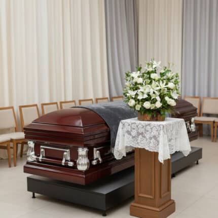 Ataúd de madera barnizada con herrajes plateados en una sala de velatorio, junto a un pedestal con mantel de encaje y un arreglo de flores blancas