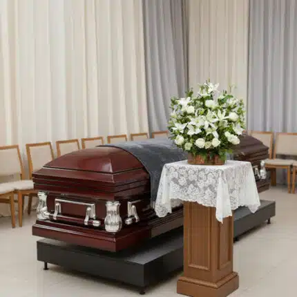 Ataúd de madera barnizada con herrajes plateados en una sala de velatorio, junto a un pedestal con mantel de encaje y un arreglo de flores blancas