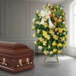 Ataúd de madera barnizada junto a corona funeraria alta con rosas amarillas, blancas y naranjas en interior iluminado