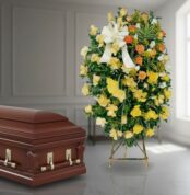 Ataúd de madera barnizada junto a corona funeraria alta con rosas amarillas, blancas y naranjas en interior iluminado