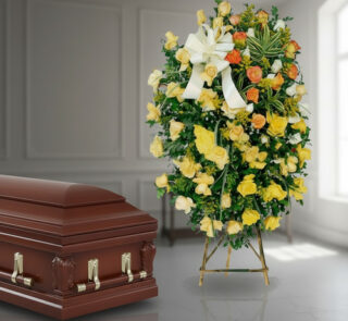 Ataúd de madera barnizada junto a corona funeraria alta con rosas amarillas, blancas y naranjas en interior iluminado