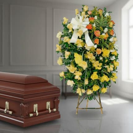 Ataúd de madera barnizada junto a corona funeraria alta con rosas amarillas, blancas y naranjas en interior iluminado