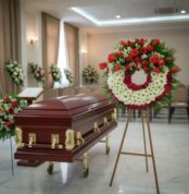 Ataúd de madera barnizada con herrajes dorados junto a una corona fúnebre de flores rojas y blancas en una sala de velatorio elegante
