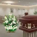 Ataúd de madera barnizada con herrajes dorados junto a un gran arreglo floral blanco y verde en una sala de velación iluminada