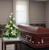 Ataúd de madera barnizada con asas metálicas junto a un arreglo floral de lirios y rosas blancas sobre una mesa en una sala de velatorio