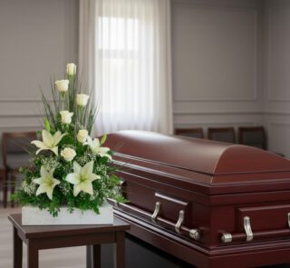 Ataúd de madera barnizada con asas metálicas junto a un arreglo floral de lirios y rosas blancas sobre una mesa en una sala de velatorio