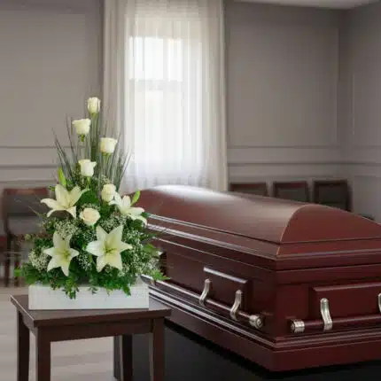 Ataúd de madera barnizada con asas metálicas junto a un arreglo floral de lirios y rosas blancas sobre una mesa en una sala de velatorio