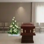 Ataúd de madera barnizada con herrajes dorados junto a un gran arreglo de flores blancas en una sala de velatorio con sillas alineadas y cortinas claras