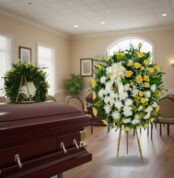 Corona fúnebre grande de flores blancas y amarillas con lazo blanco junto a ataúd de madera en sala de velatorio